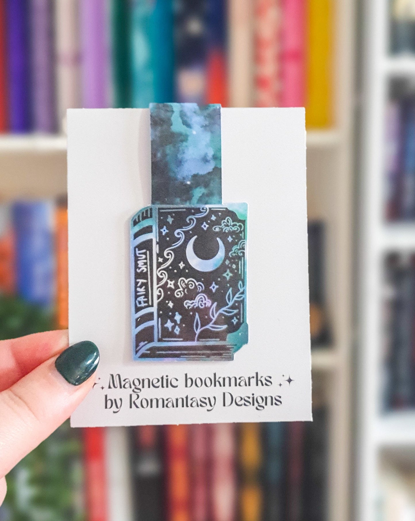 magnetic bookmark in the shape of a book with white and green clouds, stars, vines and moons on the book. The spine says fairy smut.