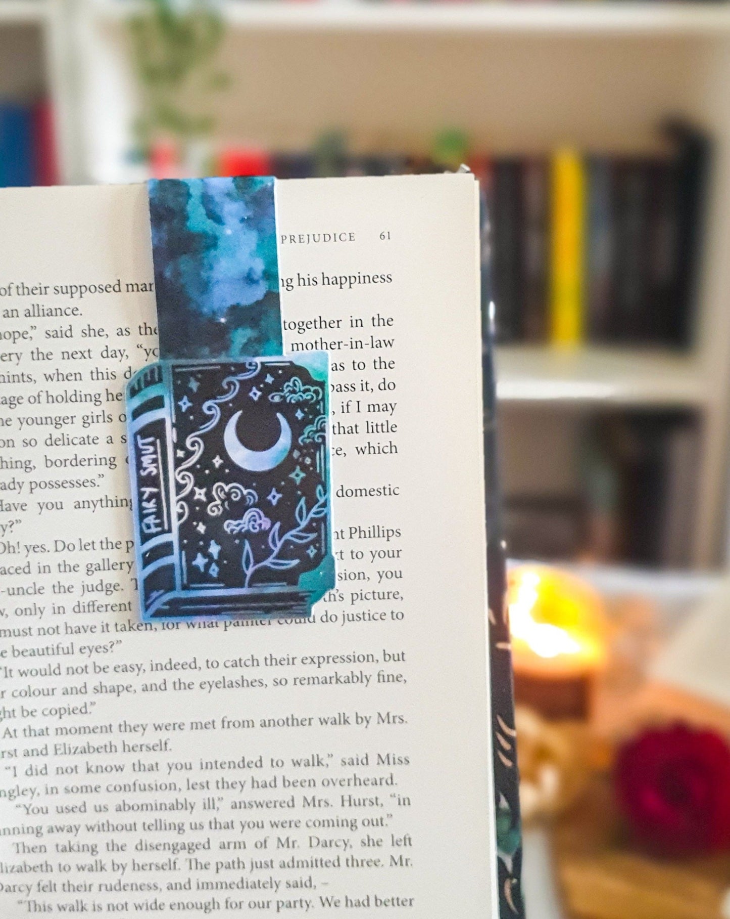 magnetic bookmark in the shape of a book with white and green clouds, stars, vines and moons on the book. The spine says fairy smut.
