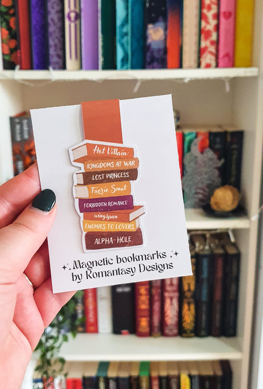 magnetic bookmark in the shape of a book stack. The spines say hot villain, kingdoms at war, lost princess, faerie smut, forbidden romance, wingspan, enemies to lovers and alpha hole. the colours are orange, brown and white.