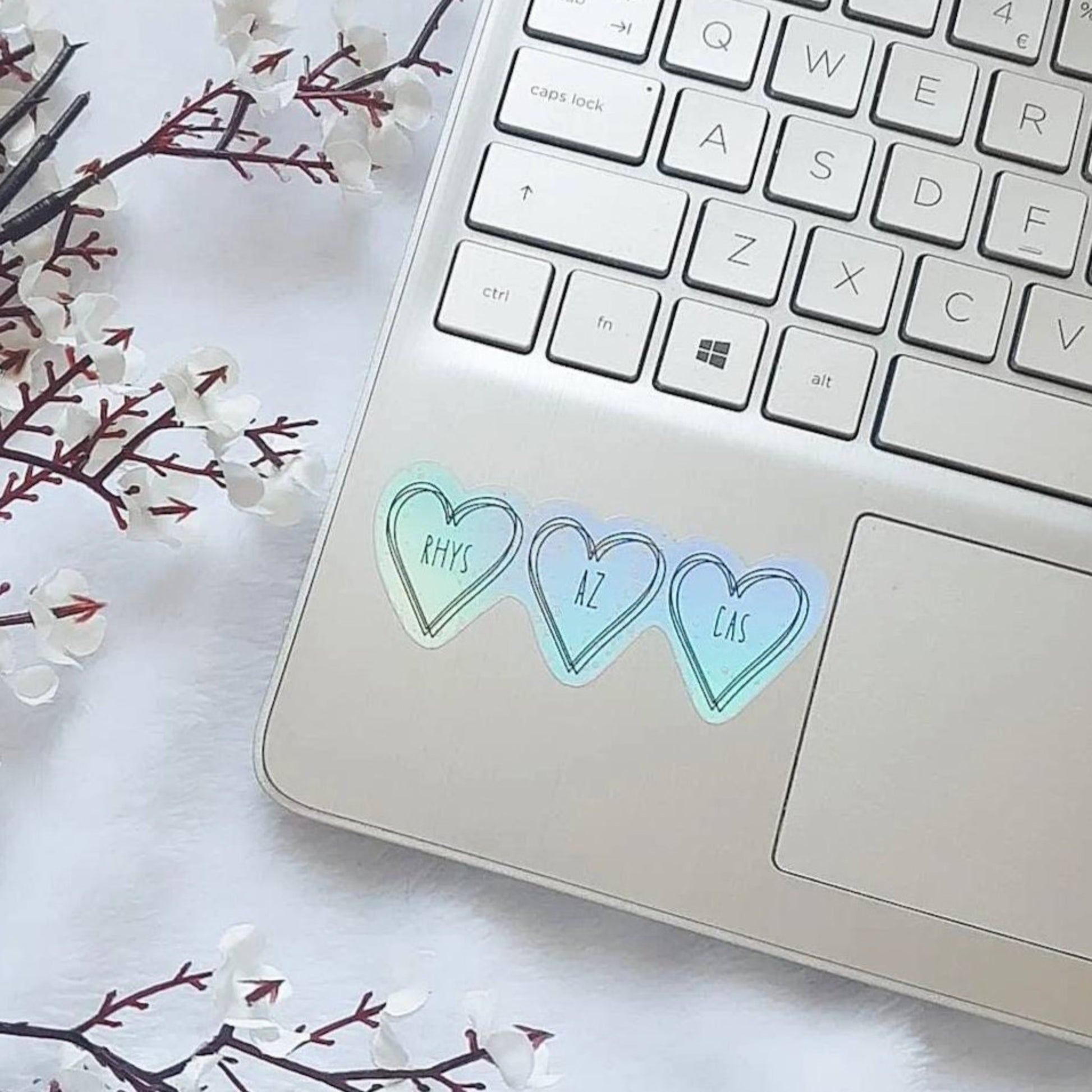 a holographic sticker with three hearts. Inside the hearts says Rhys Az and Cas