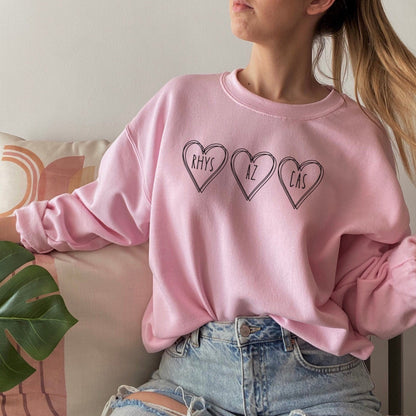 A woman wearing a pink sweatshirt and jeans. On the sweatshirt are three black heart outlines. Inside the hearts it says Rhys, Az, Cas